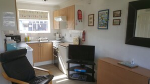 A compact kitchen with wooden cabinets, a microwave, a toaster, and a sink. There is a chair, a television on a stand, and a fire extinguisher on the wall.
