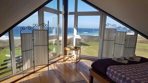 Un dormitorio con un amplio ventanal con vistas al océano, suelo de madera y una cama con colcha de diseño morado y blanco.