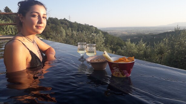 Uma pessoa relaxando à beira da piscina, com uma mesa posta com petiscos e bebidas, apreciando uma paisagem deslumbrante.