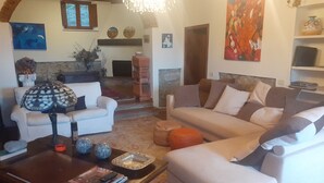 Uma sala de estar com sofá, poltronas, mesa de centro e lareira. Há quadros nas paredes e um lustre sobre a lareira.