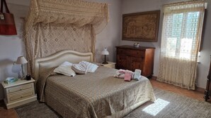 Quarto com cama, mesas de cabeceira, cômoda, janela com cortinas de renda e um quadro na parede.
