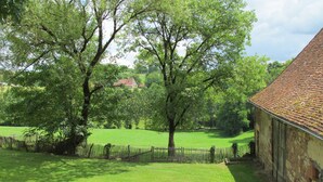 Un paysage rural avec un bâtiment en pierre, une zone délimitée par une clôture et un grand arbre.