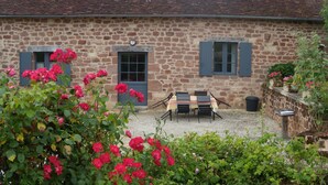 Un bâtiment en pierre avec une terrasse en gravier, une table en bois avec des chaises et des plantes en pot.