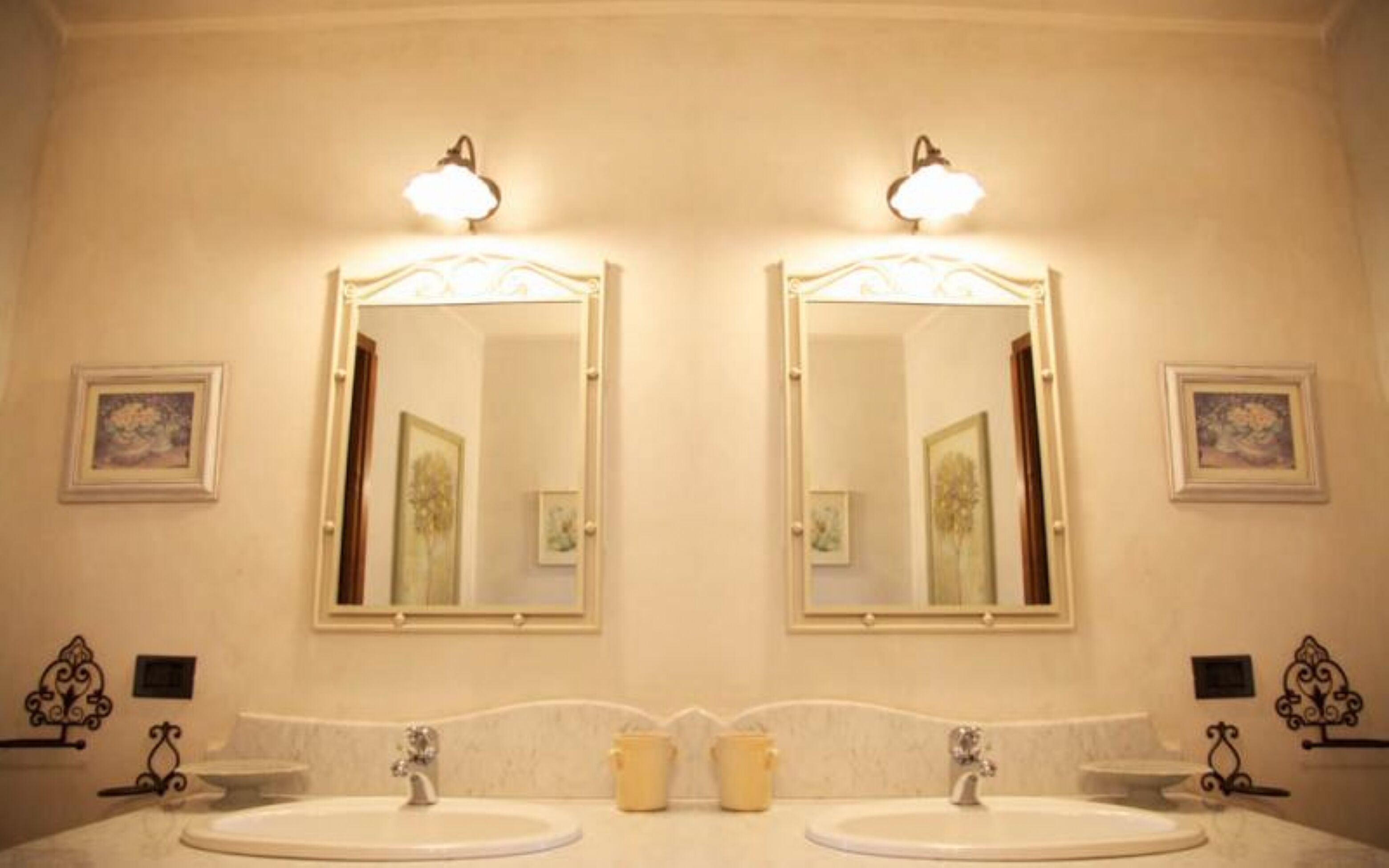 Ein Badezimmer mit zwei Waschbecken, jeweils mit einem Spiegel darüber. Über den Spiegeln sind Wandleuchten angebracht, und an den Wänden befinden sich Bilder in Rahmen.