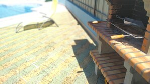 Een bakstenen barbecue met schoorsteen, een zwembad met betegeld zonnedek, en een groene constructie op de achtergrond.