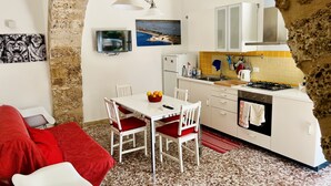 Cocina compacta con armarios blancos, estufa y lavamanos. Hay una mesa de comedor con dos sillas y un cojín rojo. Un televisor está montado en la pared sobre la mesa.