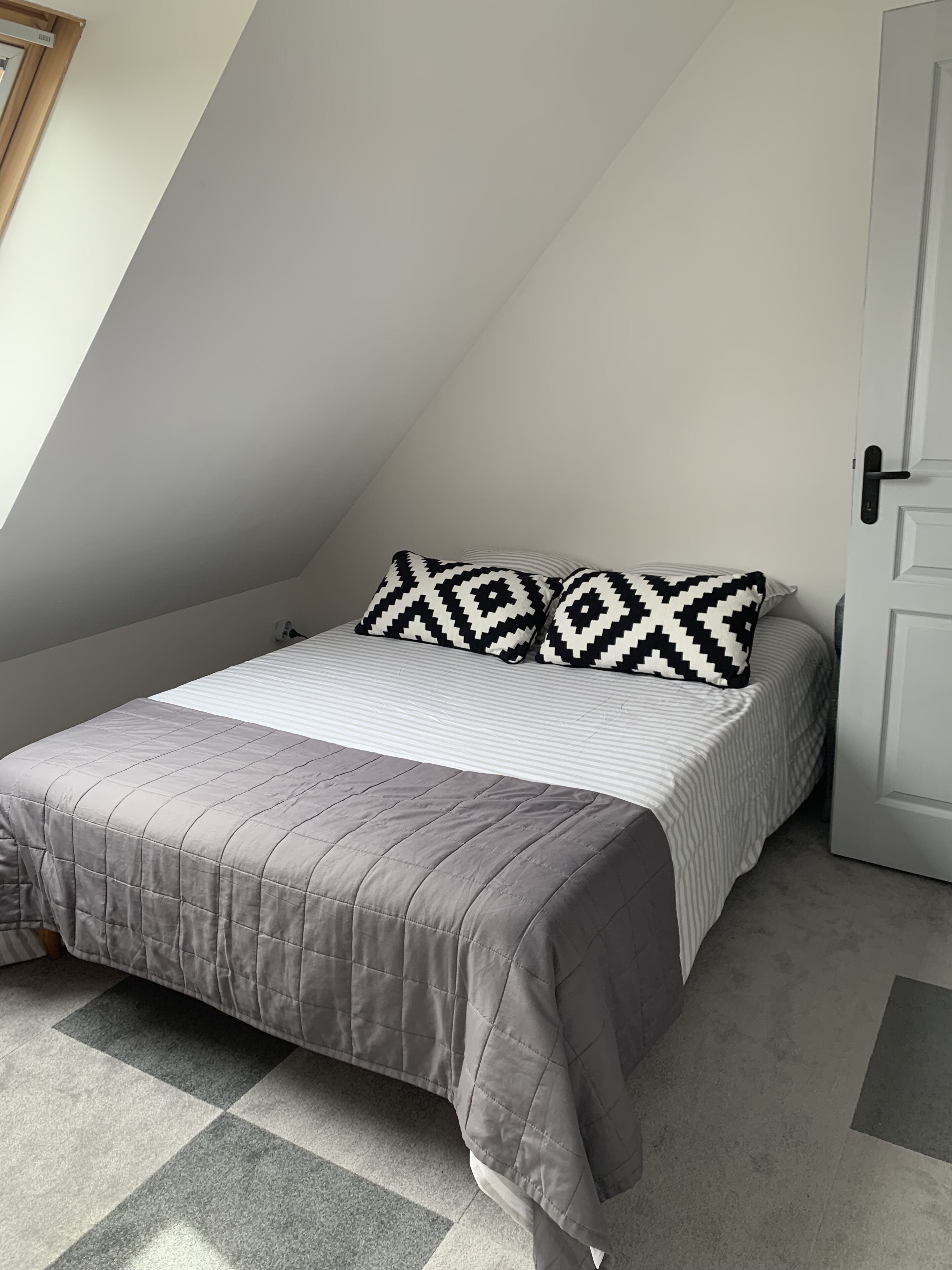 A small, neatly arranged bedroom with a single bed, a patterned pillow, and a door to the right.