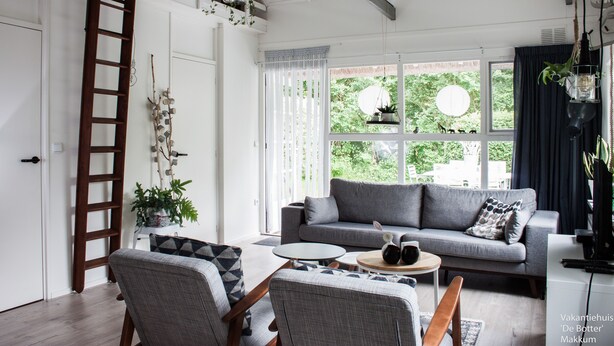 Ein modernes Wohnzimmer mit einem grauen Sofa, zwei Sesseln, einem Couchtisch und einer Holzleiter an der Wand.