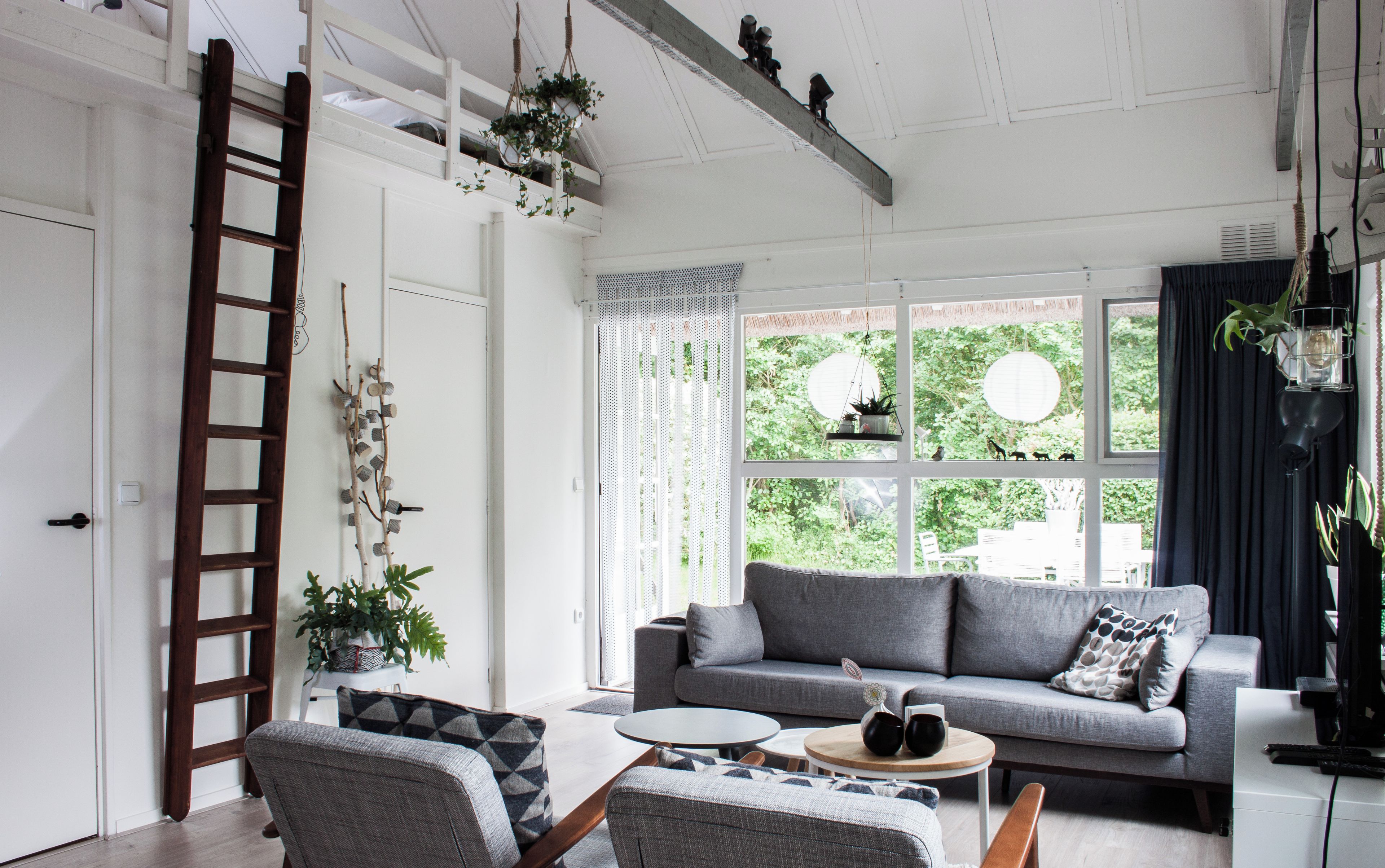 Una sala de estar moderna con un sofá gris, dos sillones, una mesita de centro y una escalera de madera apoyada contra la pared.