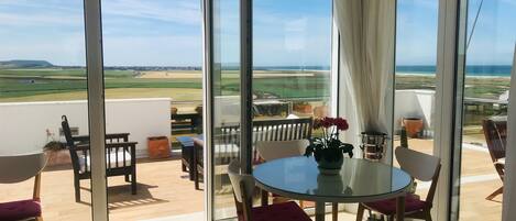 A room with a glass table and chairs, a vase with flowers, and a view of a beach and fields through large windows.