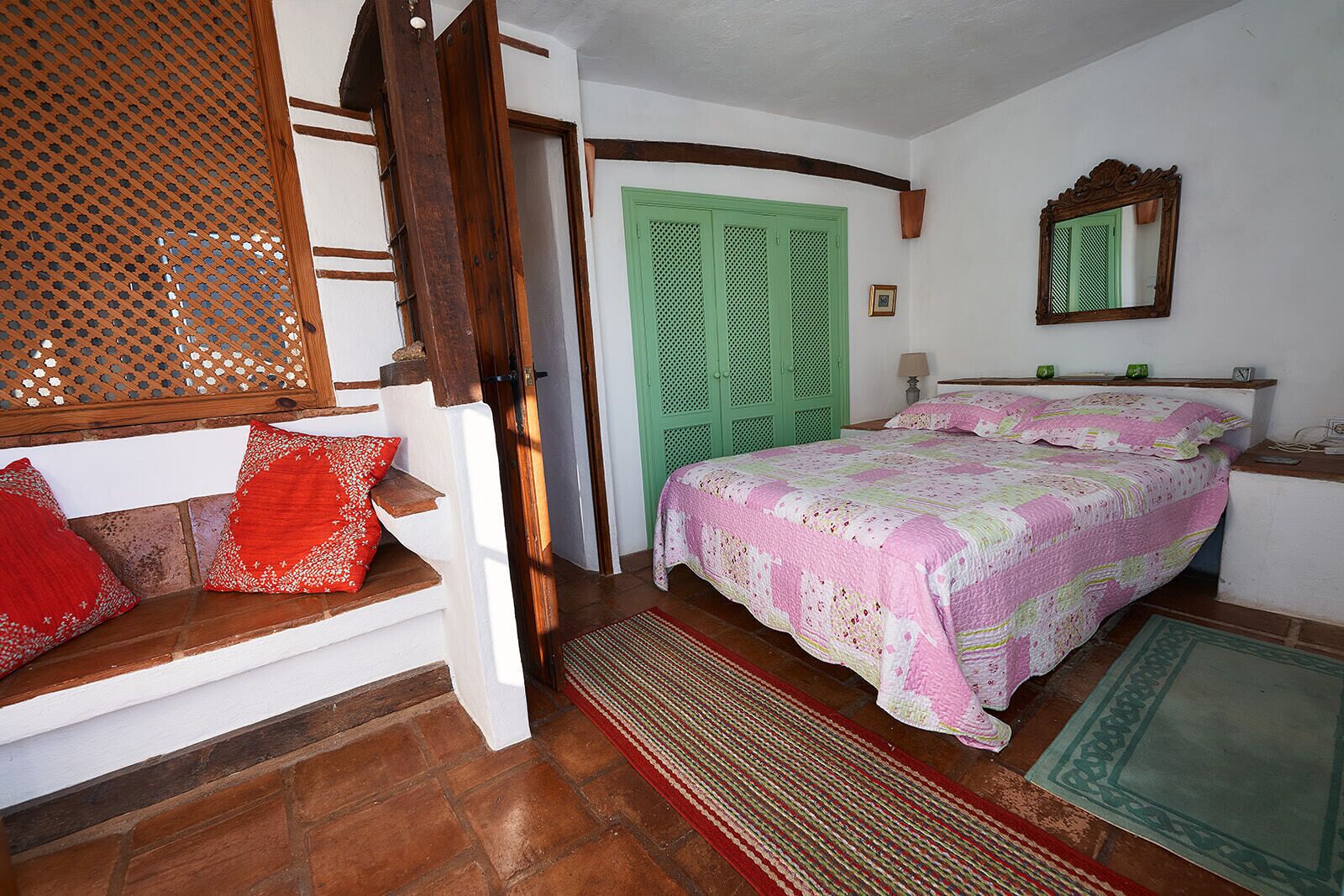 A bedroom with a wooden staircase, a green door, a bed with a pink and white quilt, and a small bedside table with a lamp.
