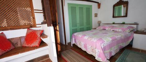 A bedroom with a wooden staircase, a green door, a bed with a pink and white quilt, and a small bedside table with a lamp.