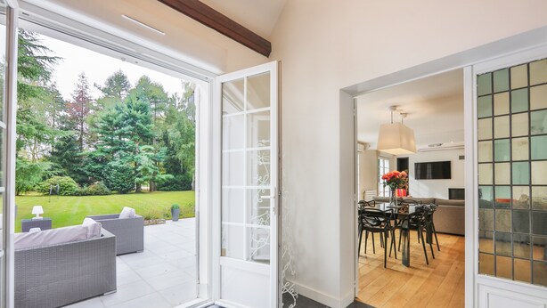 Puerta corrediza de vidrio abierta que da a un patio con mobiliario exterior y vistas a un jardín exuberante.