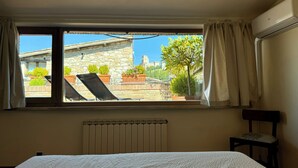 Una habitación con cama, radiador, ventana con cortinas y vista a un edificio de piedra y follaje exterior.