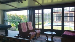 Habitació amb taula de bitlles, sofà i taula, amb finestres grans que donen vista a una piscina i vegetació a l'exterior.