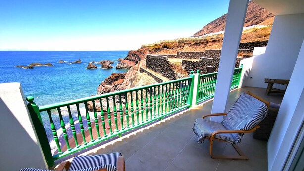 A balcony with a view of the ocean and rocky cliffs. There are two striped chairs and a wooden table.