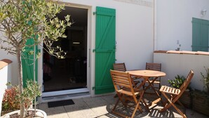 Une terrasse extérieure avec une table et des chaises en bois, une plante en pot et l’entrée d’un bâtiment avec des volets verts.