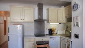 Une cuisine compacte avec des meubles de couleur claire, une hotte de cuisine en acier inoxydable, un four encastré, un lave-vaisselle et une petite table à manger recouverte d’une nappe à motifs.