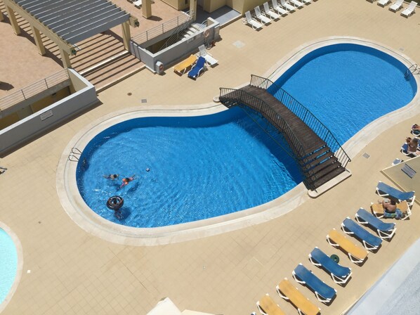 An aerial view of a swimming pool with a curved design, surrounded by lounge chairs and a small bridge.
