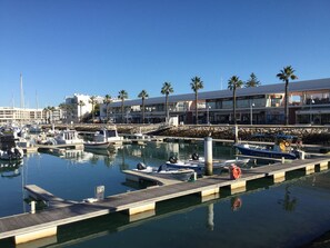 A marina with boats docked, a wooden pier, and a building with shops in the background.
