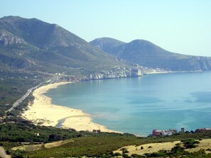 Paesaggio costiero con spiaggia sabbiosa, mare blu limpido e rilievi montuosi sullo sfondo.