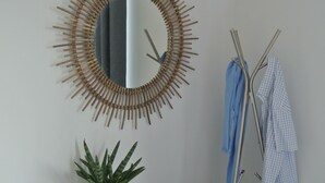 Una stanza con una poltrona azzurra, una cassettiera bianca, uno specchio con cornice in legno, una pianta e un appendiabiti con abiti.