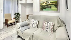 A modern living room with a beige sofa, a wooden armchair, a potted plant, and a framed landscape painting on the wall. - Suitable. Bright 10 min from the BEACH walking and 3 min from the PORT (València)
