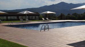 Ein Swimmingpool mit Liegestühlen und Sonnenschirmen, umgeben von einer gefliesten Terrasse und einem Gebäude mit großen Fenstern.