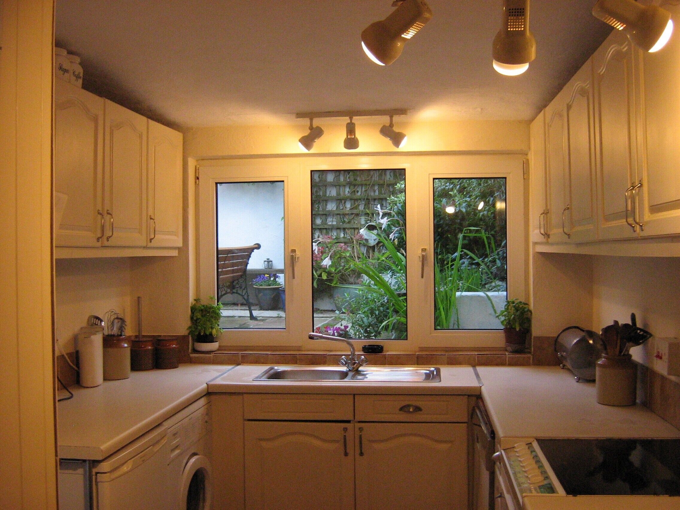 A kitchen with a built-in washing machine, a sink, and cabinets. There are two windows with views of a garden and a bench.