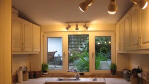 Una cocina con lavadora integrada, un lavabo y armarios. Hay dos ventanas con vistas a un jardín y un banco.