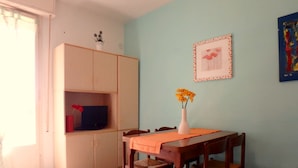 Een eetruimte met een tafel, stoelen, een vaas met bloemen, een televisie op een plank en een ingelijst beeld aan de muur.