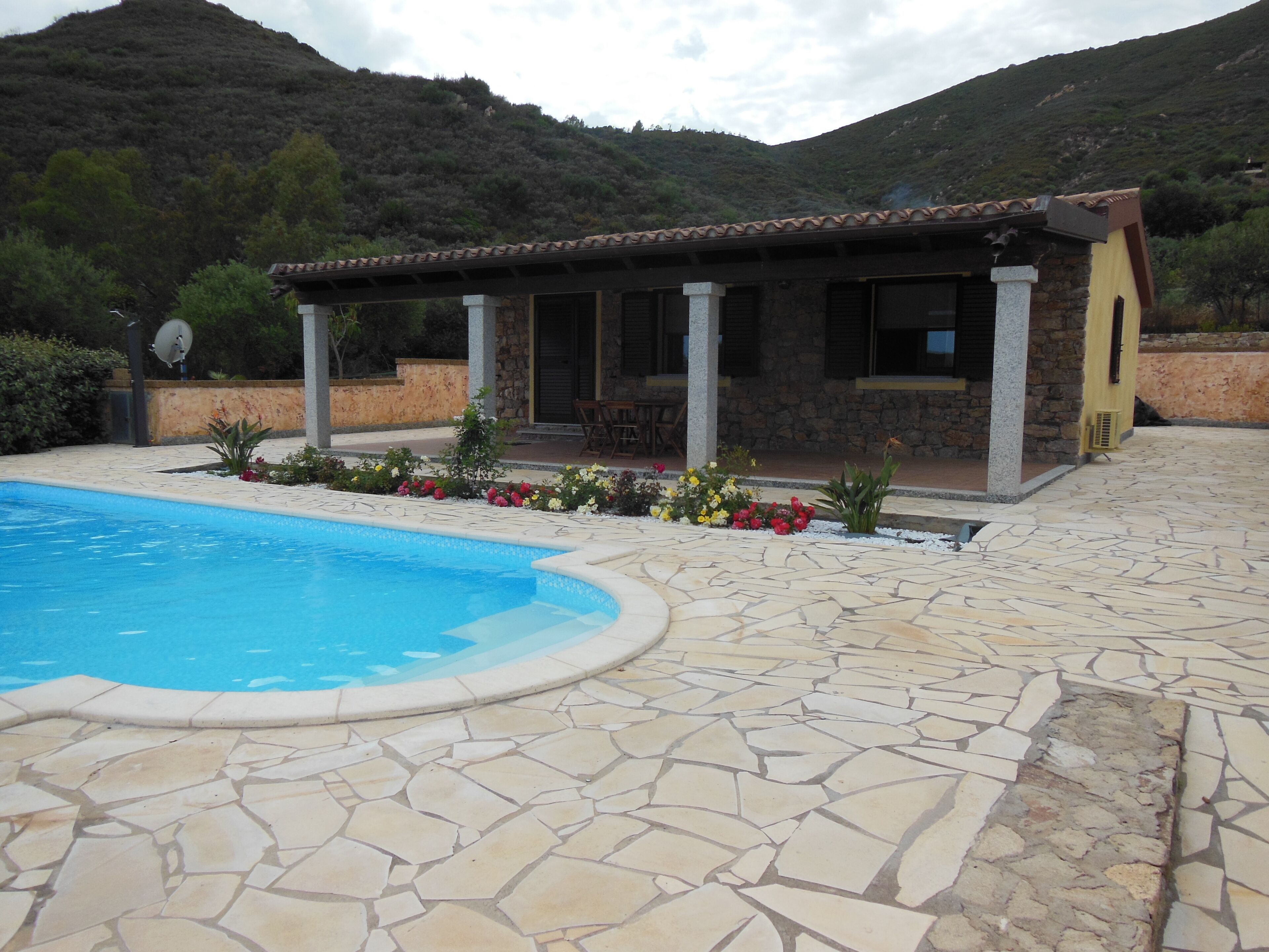 A pool area with stone flooring, a covered patio with outdoor seating, and a mountainous backdrop.