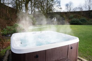 A hot tub in a garden with a stone wall and trees in the background.