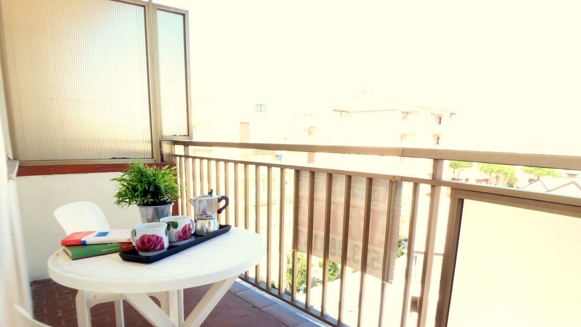 A balcony with a white table, a potted plant, a coffee maker, and a tray with cups and a book.