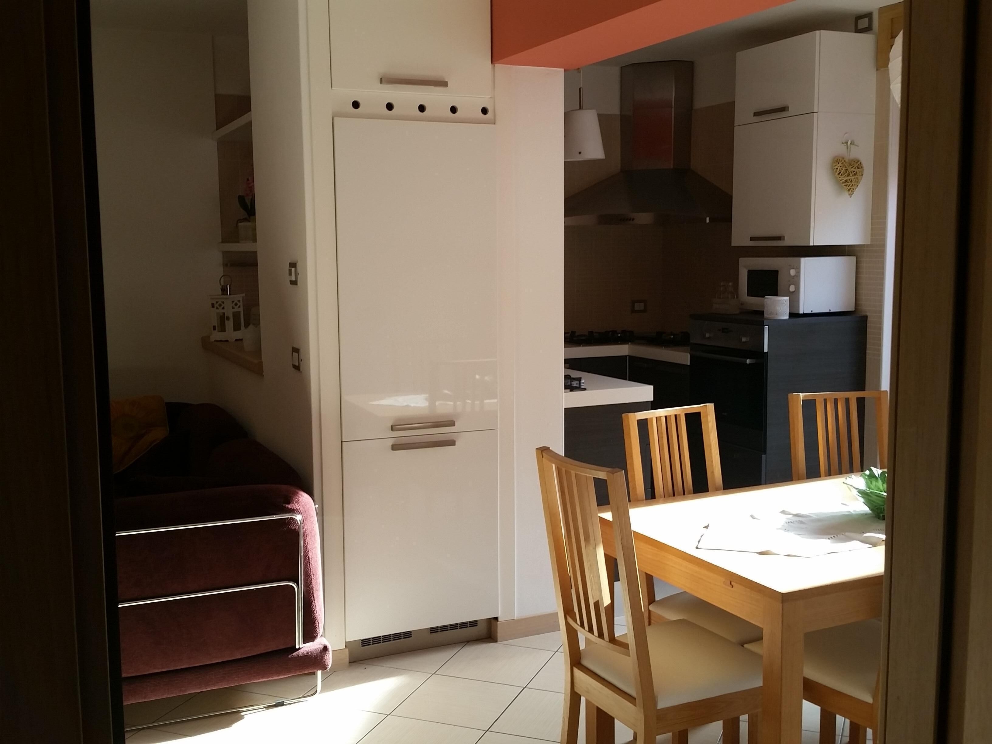 Cocina compacta con refrigerador, microondas y estufa. Hay una mesa de comedor con sillas y un pequeño rincón de descanso con sofá.