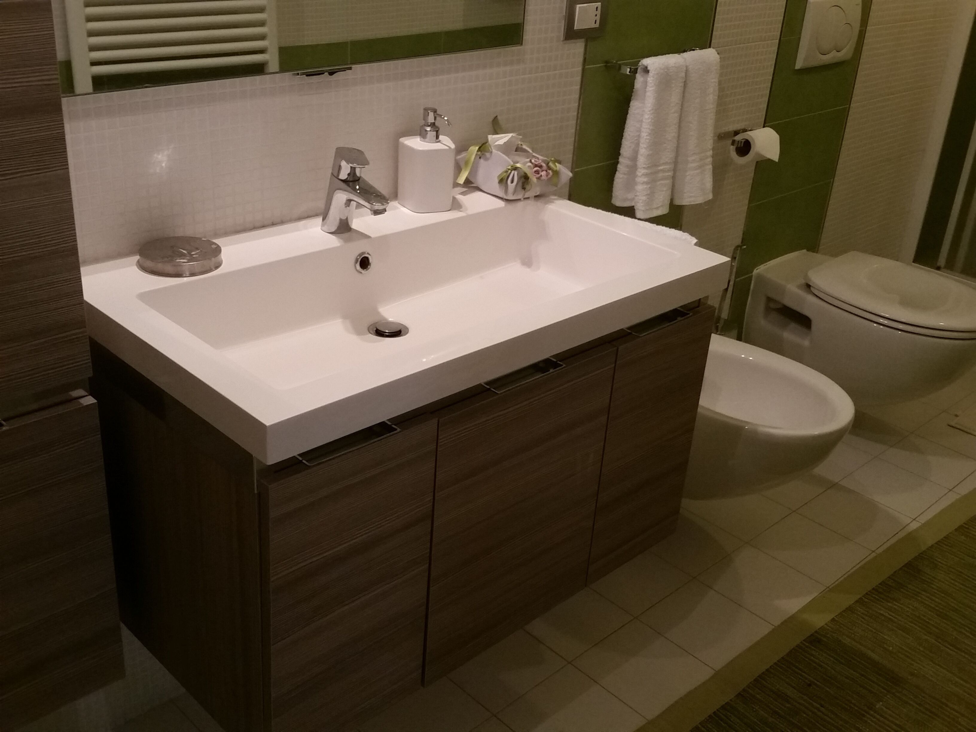 Une salle de bain moderne avec un lavabo blanc, des meubles en bois foncé, des toilettes et un mur recouvert de carreaux verts.