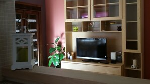 Ett vardagsrum med en platt-TV, träskåp med glasdörrar, en krukväxt och en vit, dekorativ lykta.
