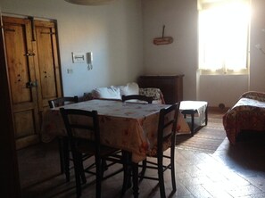 A dining area with a table and chairs, a wooden wardrobe, and a bed with a patterned bedspread.
