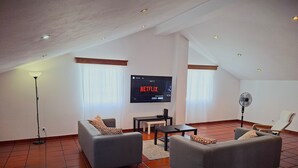 Ein modernes Wohnzimmer mit einem an der Wand montierten Flachbildfernseher, grauen Sofas mit gemusterten Kissen, einem kleinen Tisch und einem Standventilator.