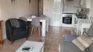 Una sala moderna con una mesita de centro blanca, un sofá gris y un sillón negro. Hay un comedor con una mesa y sillas blancas, y una cocina con armarios y electrodomésticos blancos.