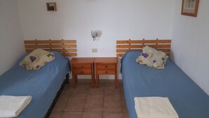 Una habitación con dos camas individuales, cada una con una colcha azul y una toalla blanca. Hay una mesita de noche de madera entre las camas, con dos cajones.