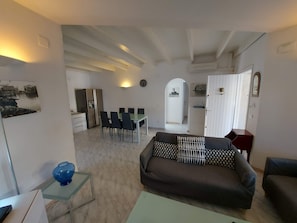 A living room with a sofa, a dining table with chairs, a kitchen area, and a hallway leading to another room.