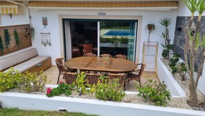 A patio with a round table and wicker chairs, surrounded by a white wall and greenery.