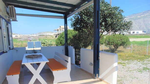 A covered patio with a table and benches, a view of the countryside, and a building in the background. - Elegant and cozy apartment (Santorini)