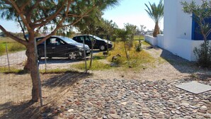 A paved driveway leading to a building with a white exterior and blue trim. There are two cars parked in the background, and a tree is visible in the foreground. - Elegant and cozy apartment (Santorini)