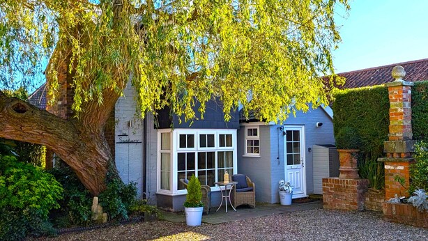 Une maison de campagne traditionnelle avec un grand arbre devant, un petit patio avec une table et des chaises, et un mur de briques avec un portail.