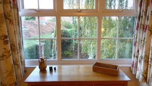 Une pièce dotée d’une grande fenêtre donnant sur un jardin, une table en bois avec un vase, un salier et un poivrier, et un panier en osier.