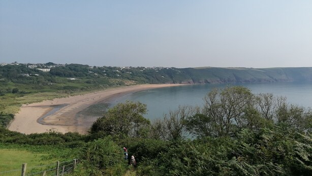 Un paisaje costero con una playa arenosa, cielo despejado y vista a un pueblo lejano.