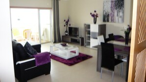 Un salon moderne avec un canapé, une table basse et un téléviseur. Une porte-fenêtre coulissante donne accès à un espace extérieur aménagé avec des chaises.