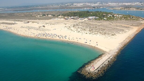 Vue aérienne d’une zone côtière avec une plage de sable, un petit quai et une mer d’un bleu limpide.